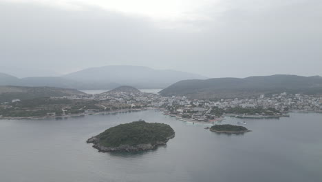 Disparo-De-Un-Dron-Volando-Sobre-El-Agua-Hacia-Las-Pequeñas-Islas-Frente-A-Ksamil-Albania-Por-La-Mañana-En-Un-Día-Gris-Nublado-Con-Barcos-Alrededor-Y-Neblina-En-El-Cielo