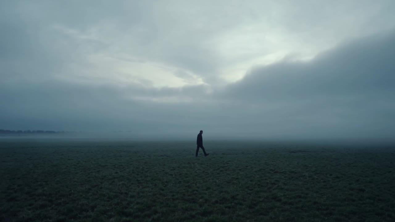 Starting to walk lone male figure wearing dark coat across misty grass plain toward right horizon