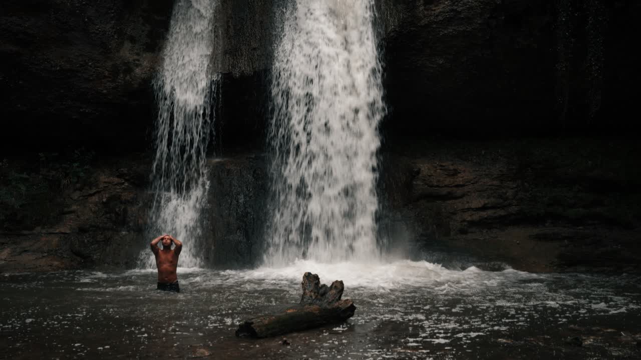 A man stands in the water in front of a waterfall, washing himself. He is topless. The natural landscape is epic. He stretches his arms toward the sky. Shot in a mystical forest in slow motion.