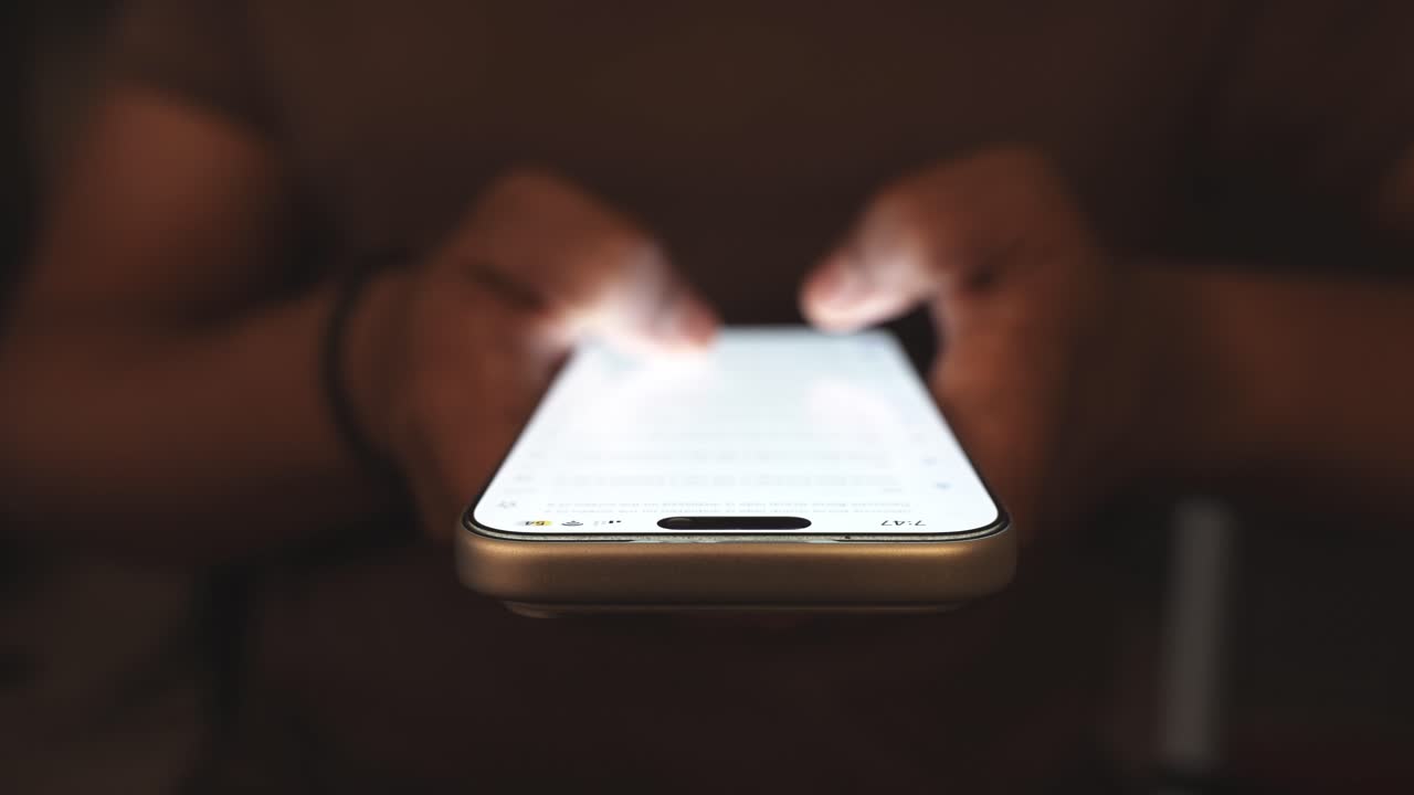 Mano che tiene uno smartphone e scorre un feed di social media in una stanza buia con una scrivania sullo sfondo, illuminata da una luce dai toni caldi. Dipendenza digitale e consumo sconsiderato di contenuti.