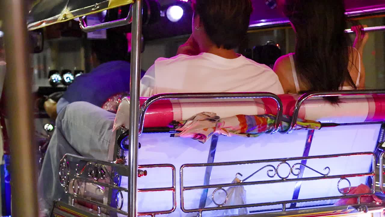 Visione posteriore di una coppia in tuk tuk che guida attraverso una strada cittadina colorata al neon di notte