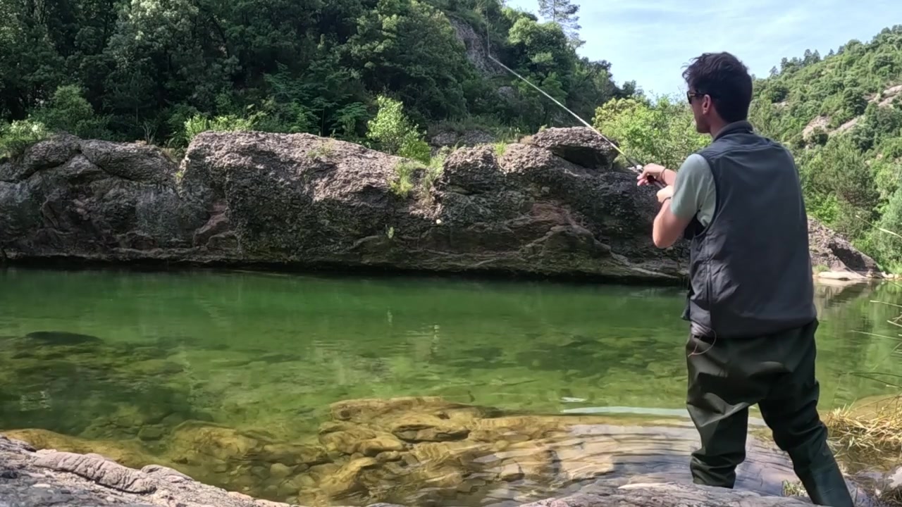 Static view of an angler standing in shallow water and casting a rod toward rocks across a clear river pool while fishing for trout and predatory fish.