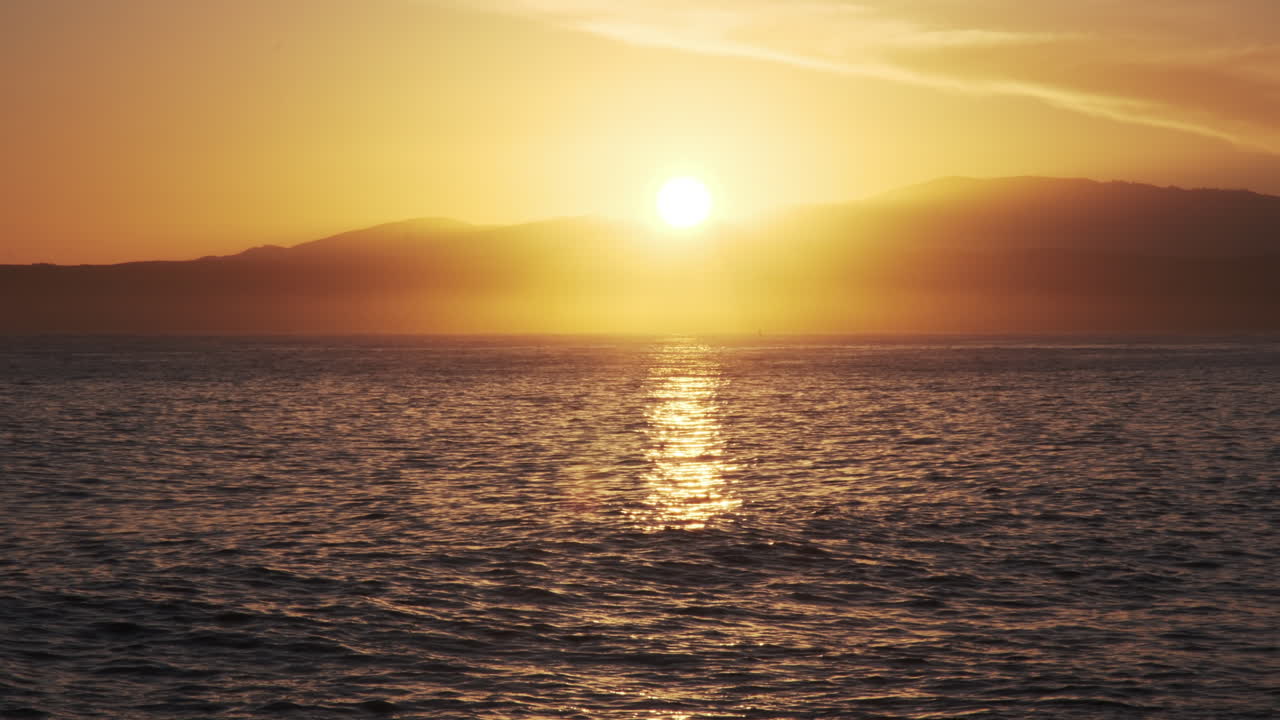 Breathtaking sunset illuminates the horizon, casting a golden glow over the ocean with distant mountains silhouetted against the bright sky