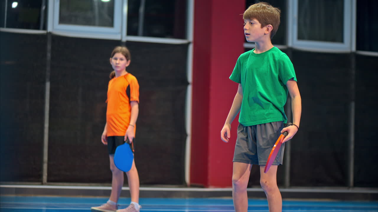 A boy and a girl training to play pickleball on a blue, inside court