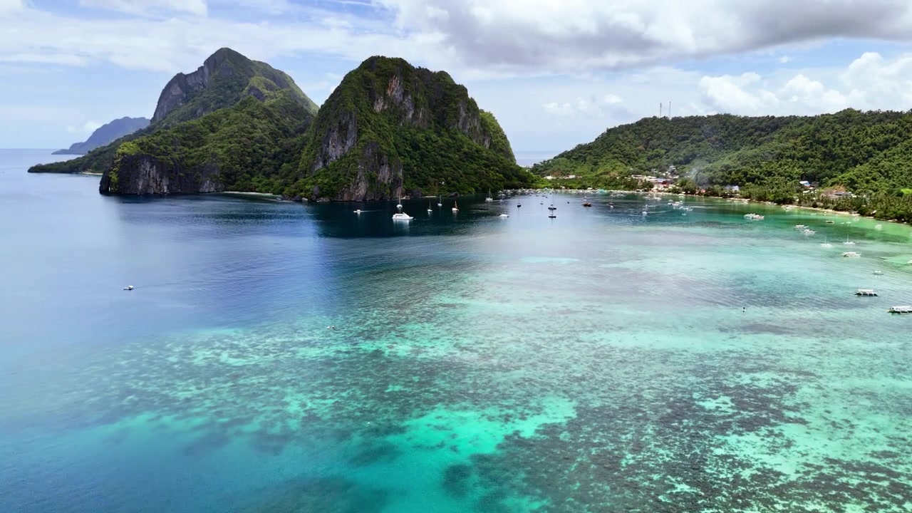 Aerial drone view of El Nido bay with two large jungle-draped limestone karst peaks, anchored sailboats and the town waterfront in turquoise water, El Nido, Palawan, Philippines.