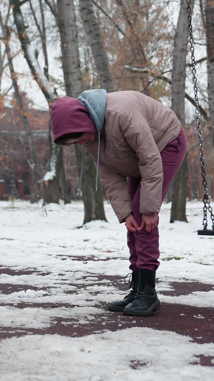 겨울 옷을 입은 여자가 무을 구부리고 을 때 무에 통증이 느지며, 벌거벗은 나무와 눈으로 인 땅의 배경으로 눈이 많은 공원에서 천천히 흔들리고 있습니다.