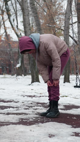 mujer en traje de invierno siente dolor en la rodilla mientras se dobla y frota la rodilla, balanceándose balanceándose lentamente en un parque nevado con fondo de árboles desnudos y suelo cubierto de nieve
