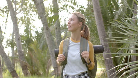 young caucasian woman hikes through a lush park, looking around with a smile