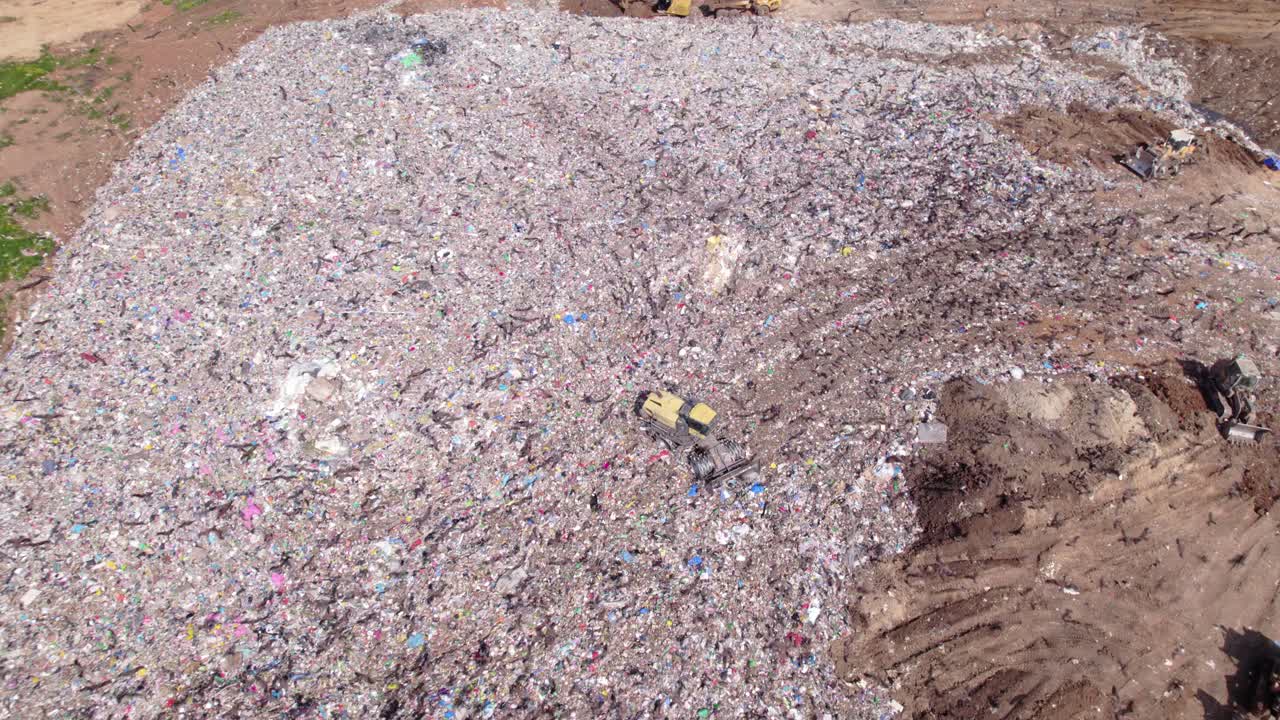 Aerial drone shot of a bulldozer operating on a large landfill mound while dozens of birds of prey circle and fly above the waste site, highlighting human impact, waste management activity, and wildli