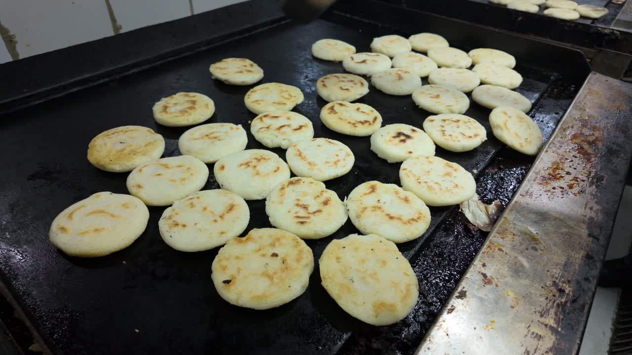 many arepas cooking on a hot, greasy commercial griddle. A hand moves them as they brown. High-volume, traditional food prep