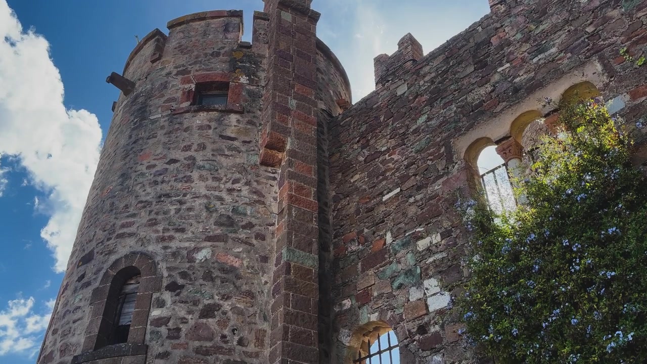 Vista dal basso di una torre di castello in pietra consumata dalle intemperie, mura merlate e finestre ad arco incorniciate da viti fiorite sotto luminose nuvole mediterranee