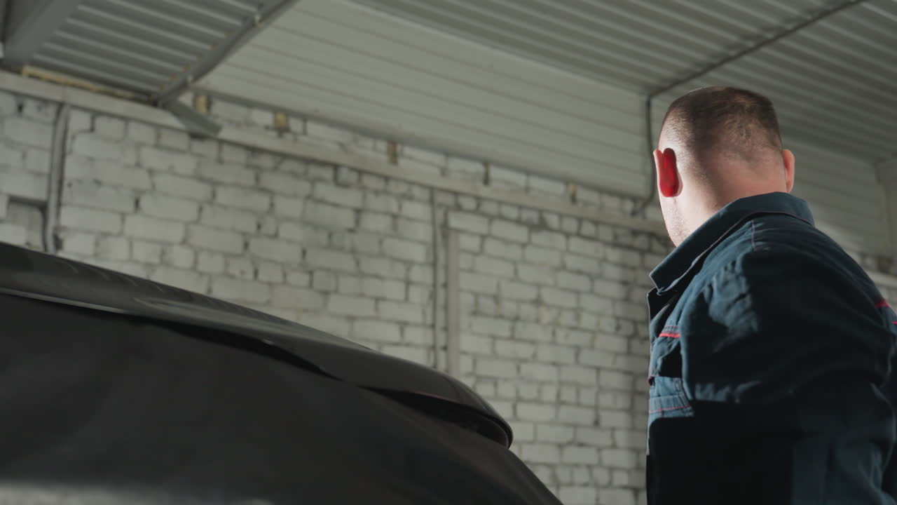 механик в синей форме надежно закрывает капот черной машины внутри механической мастерской, гарантируя, что все проверки технического обслуживания завершены