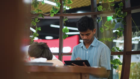 moving shot capturing white boy resting head on wooden table while friend sits behind reading tablet behind decorative pillar blocking partial view offering candid study moment under soft ambient light