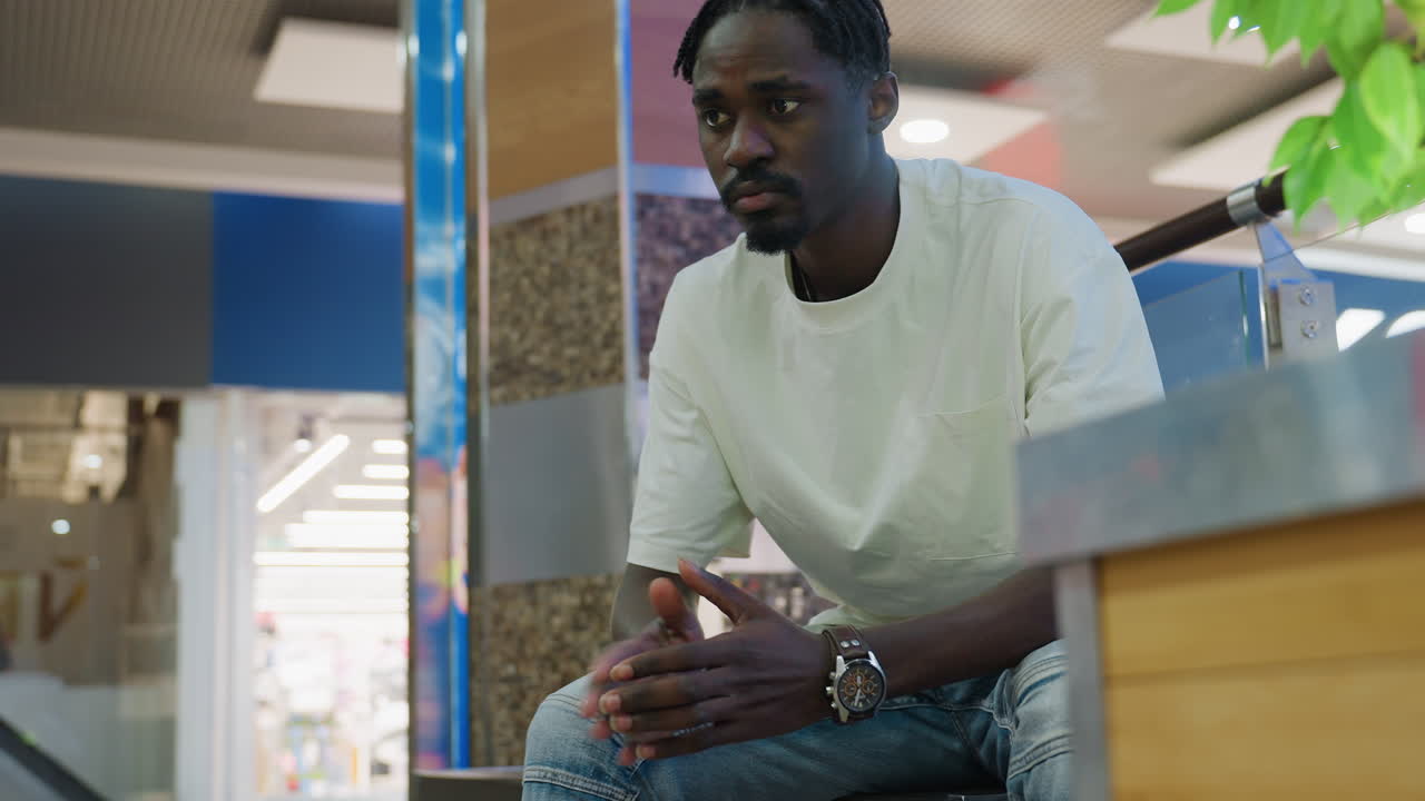 Young man in white shirt and jeans seated indoors in shopping mall tapping hands together with watch on wrist thoughtful expression waiting for someone near green plant urban lifestyle