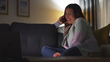 Smiling-Woman-Sitting-On-Sofa-At-Home-At-Night-Talking-On-Mobile-Phone-And-Watching-Movie-Or-Show-On-Laptop-2