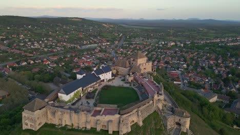 a panoramic aerial view captures a hungarian historic medieval castle perched atop a rocky hill, with its fortified walls, towers, and inner courtyard clearly visible.