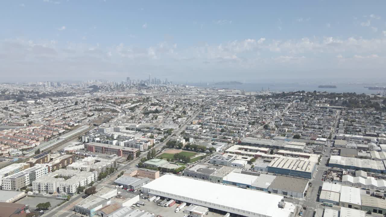 This drone shot offers a sunny, scenic vista of San Francisco's Bayview neighborhood, commercial and residential areas, downtown skyline, and Yerba Buena Island, all bustling with bay activity