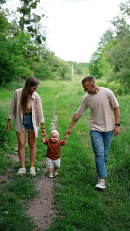 happy proud young parents holding their little baby by the hands. lovely kid taking slow steps learning to walk in the nature. vertical video.