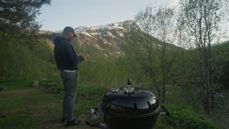 hombre abriendo cerveza mientras está de pie junto a la parrilla y disfrutando del paisaje natural