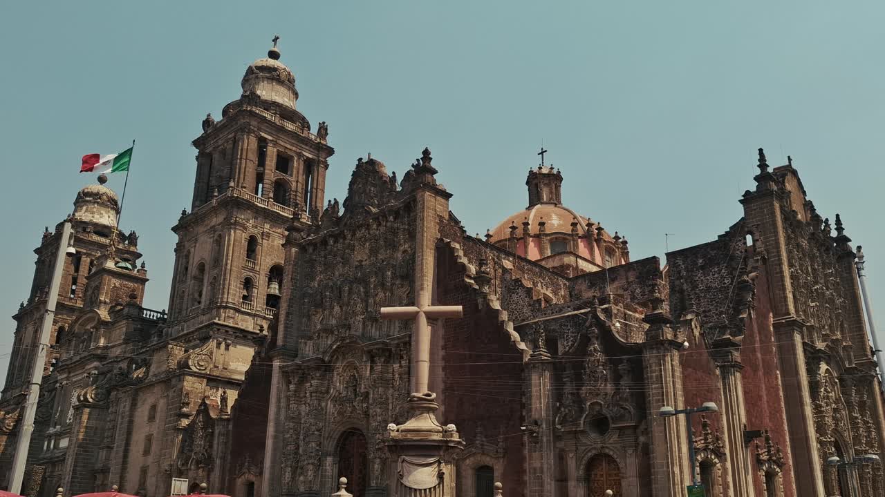 Close-up view of the ornate baroque façade and towers of the Mexico City Metropolitan Cathedral, with the Mexican flag waving in the background under a clear sky