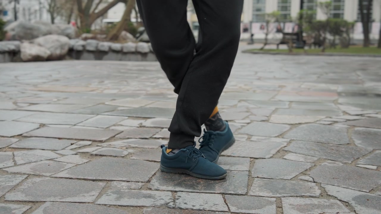 Närbild på en persons fötter i blå sneakers och svarta byxor som utför fotarbete på våt kullerstensbeläggning. Personen lyfter upp ett ben upprepade gånger, vilket visar rörelse mot den texturerade stenen.
