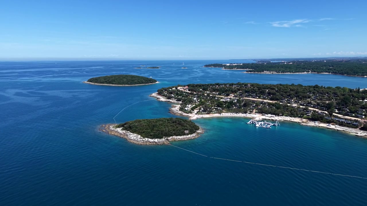 Pienet metsäiset saaret täplittävät kirkkaan sinisiä Adrianmeren vesiä Istrian rannikon edustalla, ankkuroitujen purjeveneiden ollessa hiekkarannan leirintäalueen ja mäntymetsäisen rannikon lähellä. Kroatia. Ilmakuva drone-laajakuva.