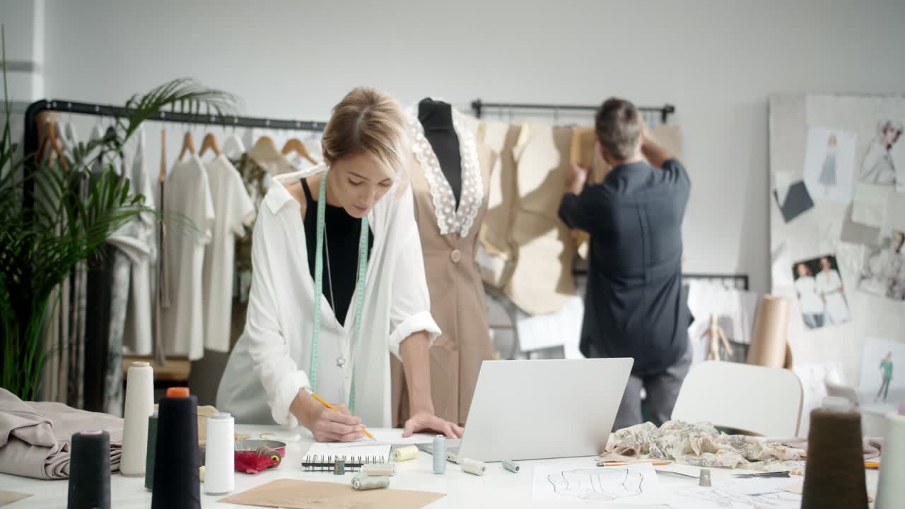 여성 재단사가 테이블에 메모를 하고, 동료 남자가 그녀에게 다가가 옷 조각을 선택합니다.