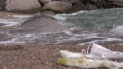 Polluted-mask-and-glove-on-a-beach-with-rushing-wave-nearby,-static-shot-focusing-background