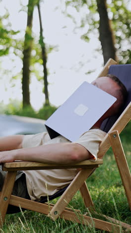 man sits in the wood in a folding chair his face covered with laptop. man smiles when the laptop descends. car at backdrop. vertical video