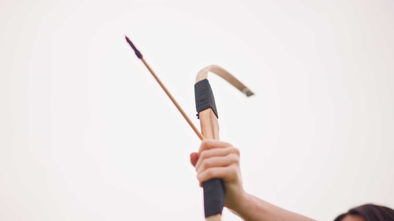 Eine weiße Frau legt einen Pfeil an und zielt mit ruhiger Hand, Nahaufnahme von Bogen und Pfeil vor blassem Himmel, Wettkampftrainingsatmosphäre, Sonnenbrille und gelassene Haltung, technikorientierter Schuss