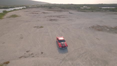 Vista-Aérea-De-Un-Viejo-Coche-Muscular-Americano-En-Un-Desierto-Durante-Una-Puesta-De-Sol-Junto-A-Un-Río