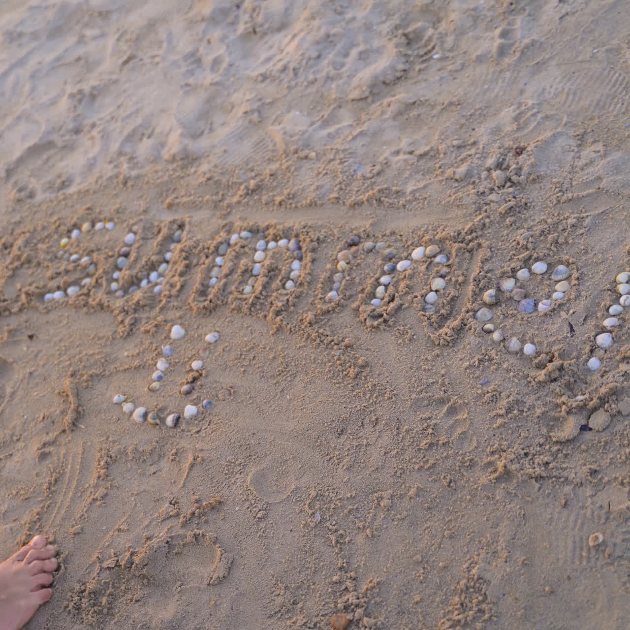 Boy is putting with pebbles the word summer on a sand beach. Child laying the smiley sign with small shells on sand background. Happy childhood.
