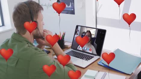 Multiple-heart-balloons-floating-against-man-wearing-headset-having-a-video-call-on-laptop-at-office