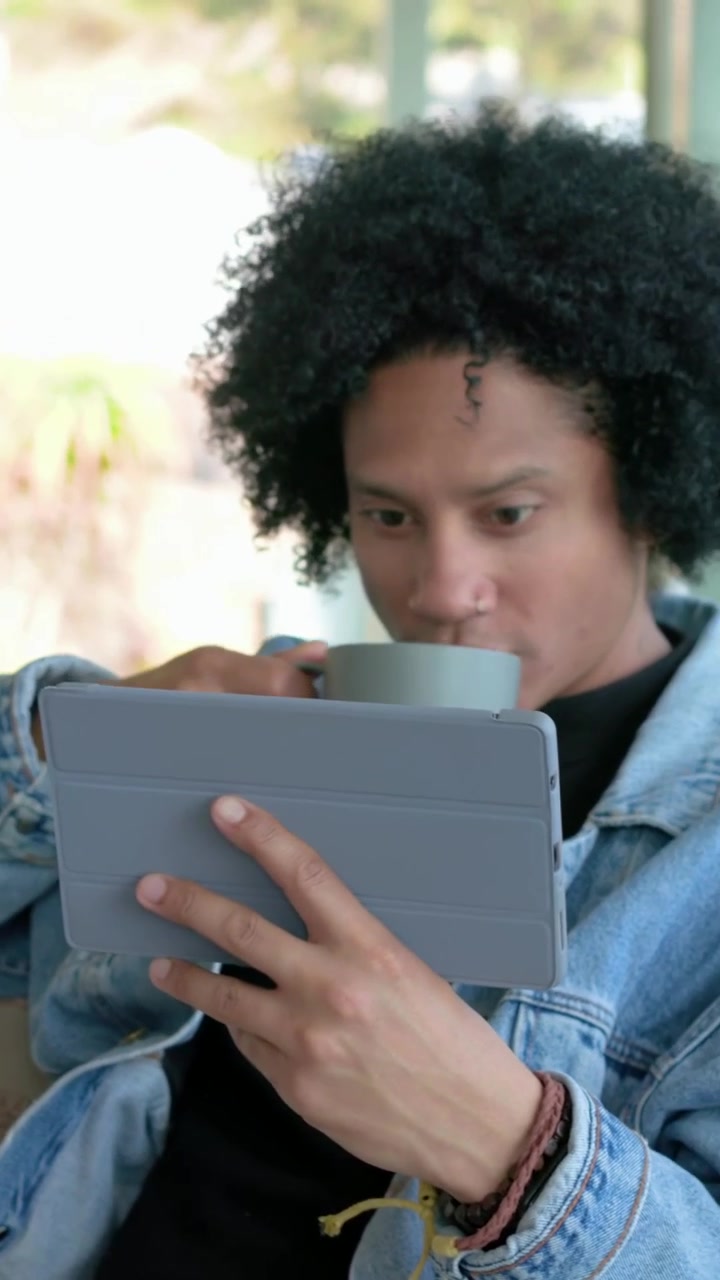 Vertical video: Reading tablet man in denim shifting on sofa by glass doors, lifting mug, sipping
