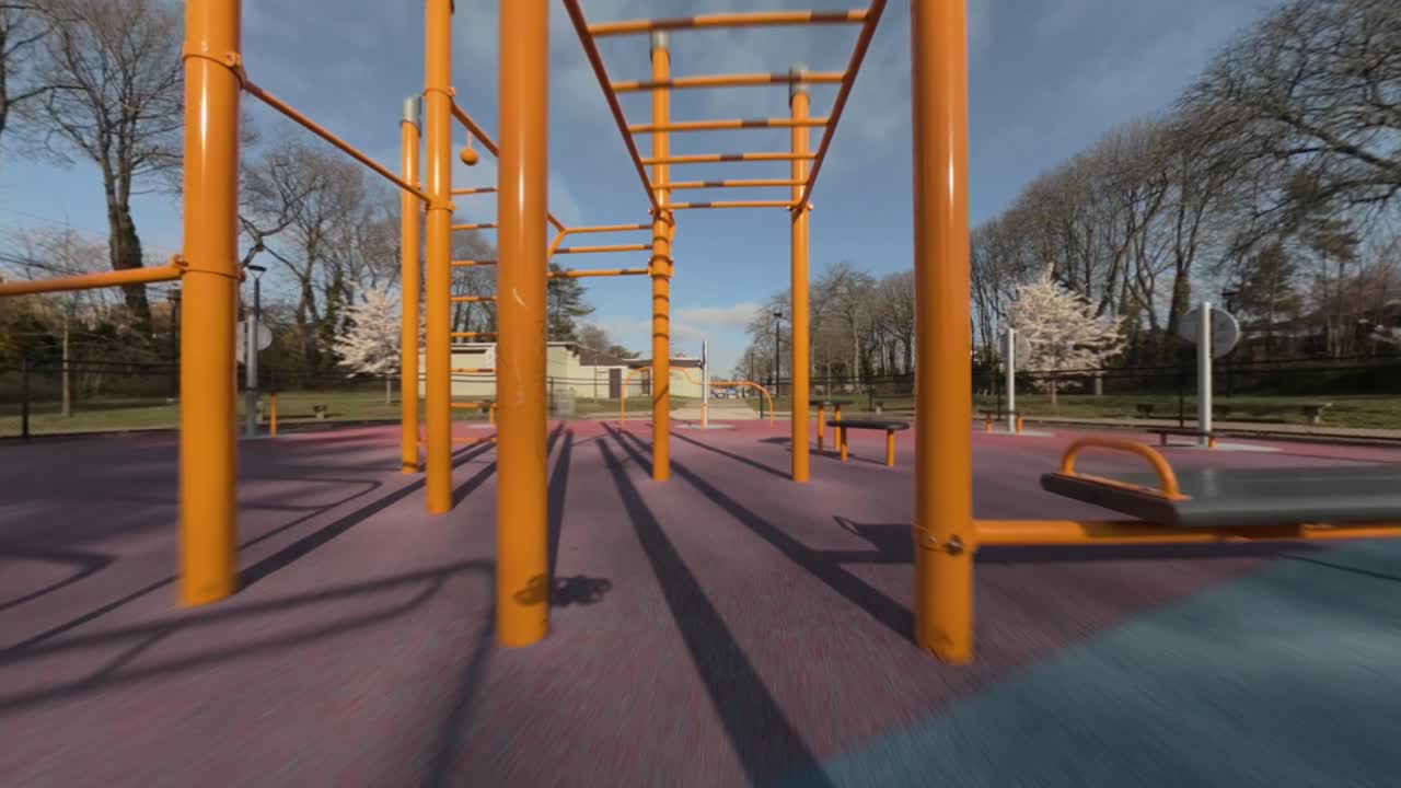 Un vuelo de dron FPV a baja altitud a través de barras de mono y una estructura de juegos doradas y amarillas en un parque infantil bajo un sol brillante, capturando un movimiento rápido y juguetón en un parque. La cámara se desplaza entre los postes.