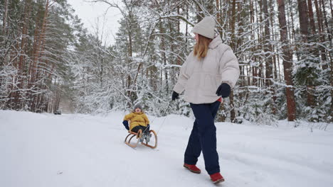 una joven madre y su hijo se divierten en invierno en el bosque en trineo a cámara lenta. mamá feliz de paseo con su hijo en un bosque nevado