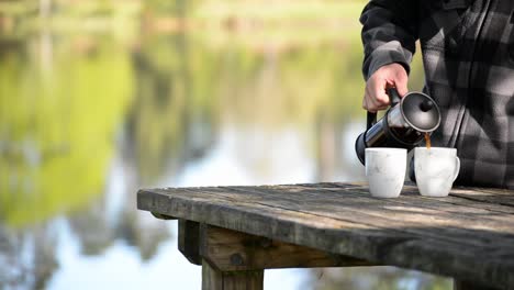 man in checked flannel pouring coffee from french press into two mugs, outdoors