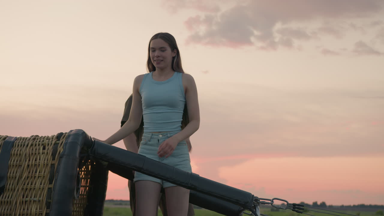 young couple climbs out of toppled wicker hot air balloon basket on grassy field after landing during vibrant sunset with visible burner system and countryside horizon in background