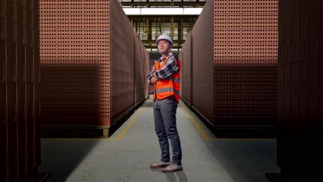 full body side view of asian male engineer with safety helmet crossing his arms and looking around while standing with red brick packed in stacks are stored