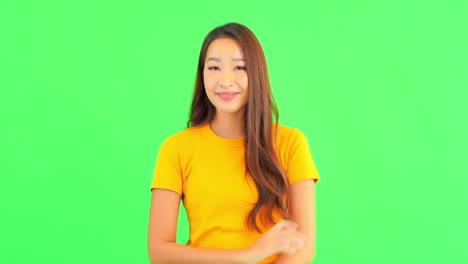 beautiful asian woman wearing yellow t-shirt crossing arms and showing ok gesture with two hands smiling looking to camera isolated, studio shot on a green studio background with copy space for text