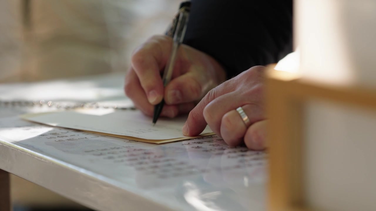 Mani di persona sposata che scrive su carta votiva alla scrivania del tempio con penna e anello visibili, Giappone