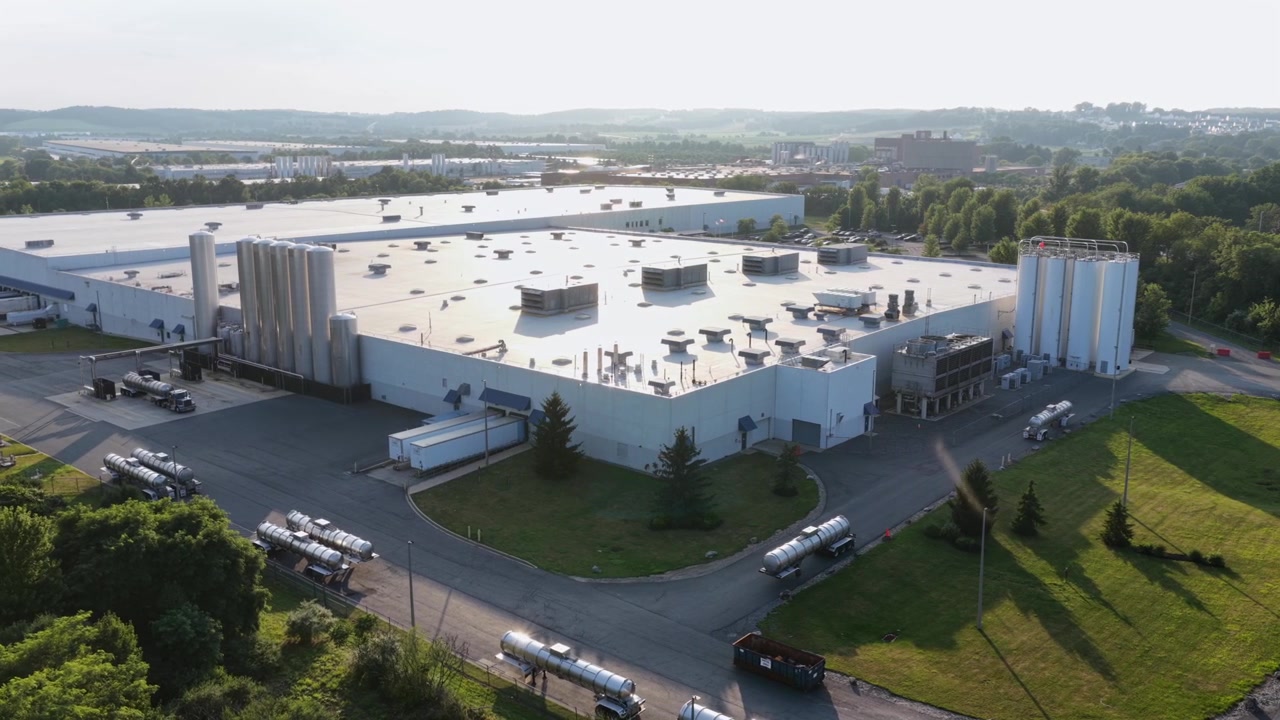 Sølvfarvede tankbiler står langs læsseramper ved siden af en massiv hvid industribygning med HVAC-enheder på taget og høje lagersiloer nær bakker i Allentown, Pennsylvania, USA. Bredt luftfoto fra drone