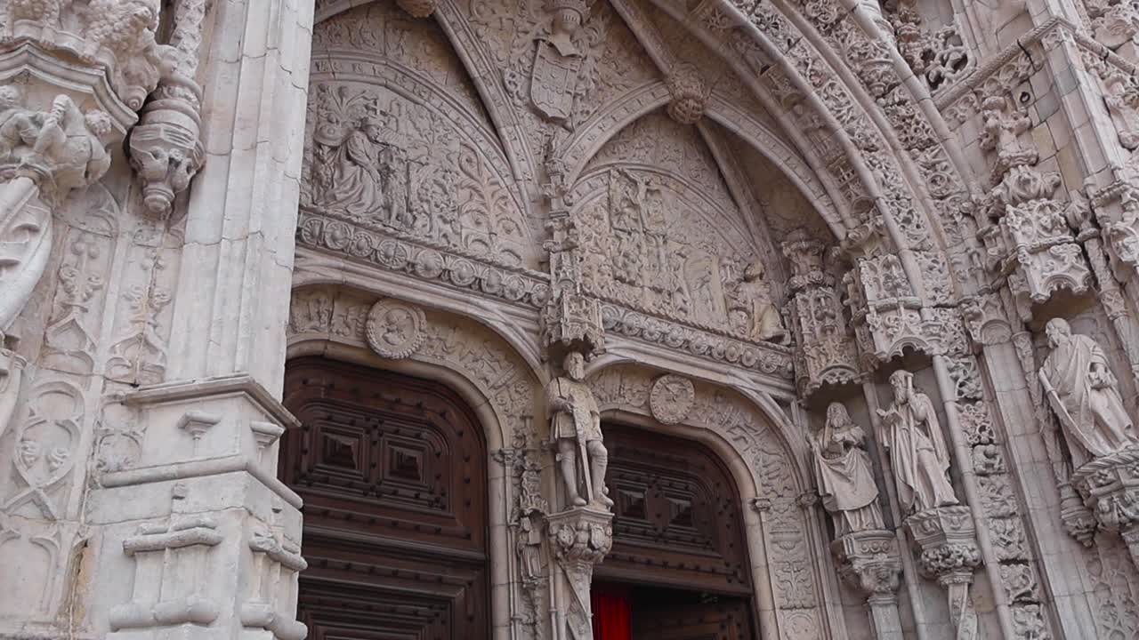 Svelata cinematografica dal basso verso l'alto dell'ingresso del Monastero dos Jerónimos che mostra sculture in pietra intagliata e porte in legno aperte con una tenda rossa all'interno di un famoso punto di riferimento di Lisbona