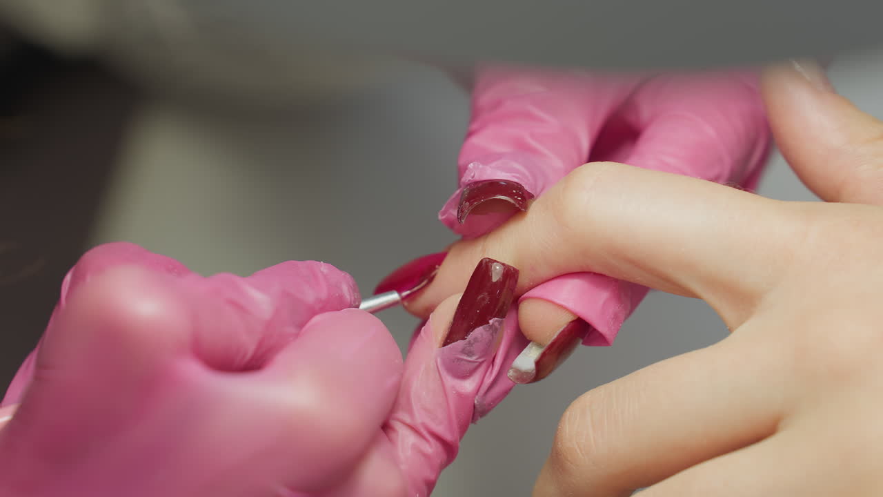 Close up of nail technician wearing pink gloves using fine brush to carefully apply red nail polish to side of client fingernail, demonstrating precision and attention during manicure procedure