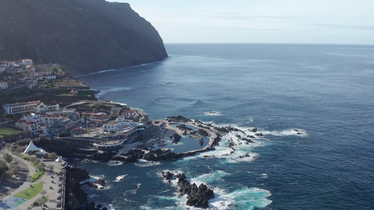 Splendida e serena città costiera affacciata su un vasto oceano, con onde che si infrangono, rocce vulcaniche nell'acqua, piscine naturali e un'imponente montagna sullo sfondo.