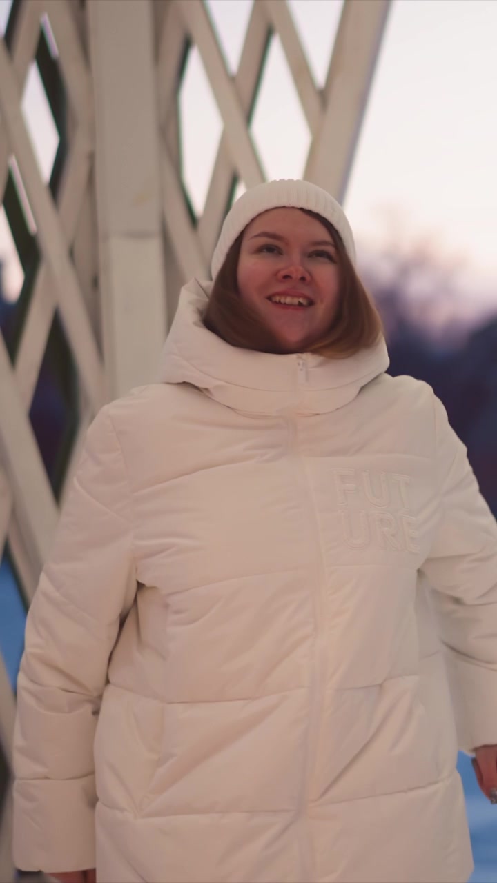 Woman in white puffer jacket and knit hat moves through outdoor winter setting adjusting her clothing as woman walks. background features soft evening light with blurred cityscape elements