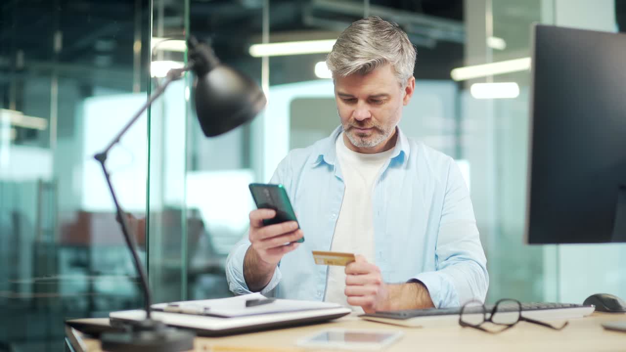 Happy man shopping online pays by credit card using mobile phone, smartphone sitting at office desk, at work. Purchase travel, bank service, booking.