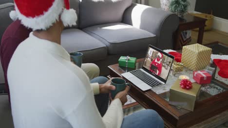 Padre-E-Hijo-Birraciales-Con-Gorros-De-Papá-Noel-Usando-Una-Computadora-Portátil-Para-Una-Videollamada-Navideña-Con-Papá-Noel-En-La-Pantalla