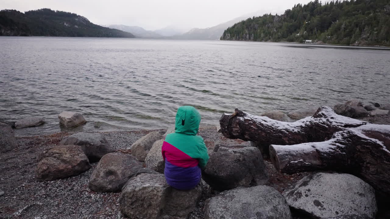 Young child in colorful jacket reflects in silence by cold Patagonian lake, emotional depth, childhood introspection