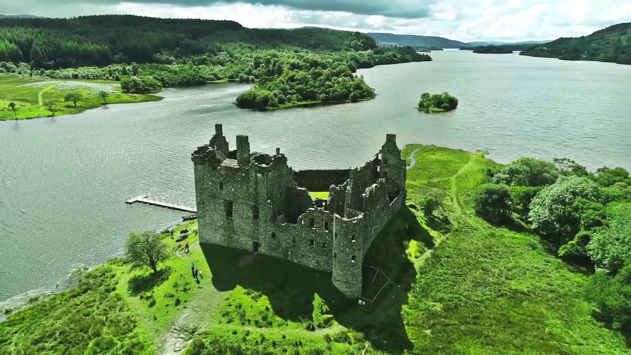Riprese aeree cinematografiche con drone che orbita lentamente attorno a un remoto castello scozzese abbandonato, circondato da un aspro paesaggio delle Highlands. La telecamera si muove elegantemente attorno alle rovine in pietra consumate dal tempo, rivelando drammatici